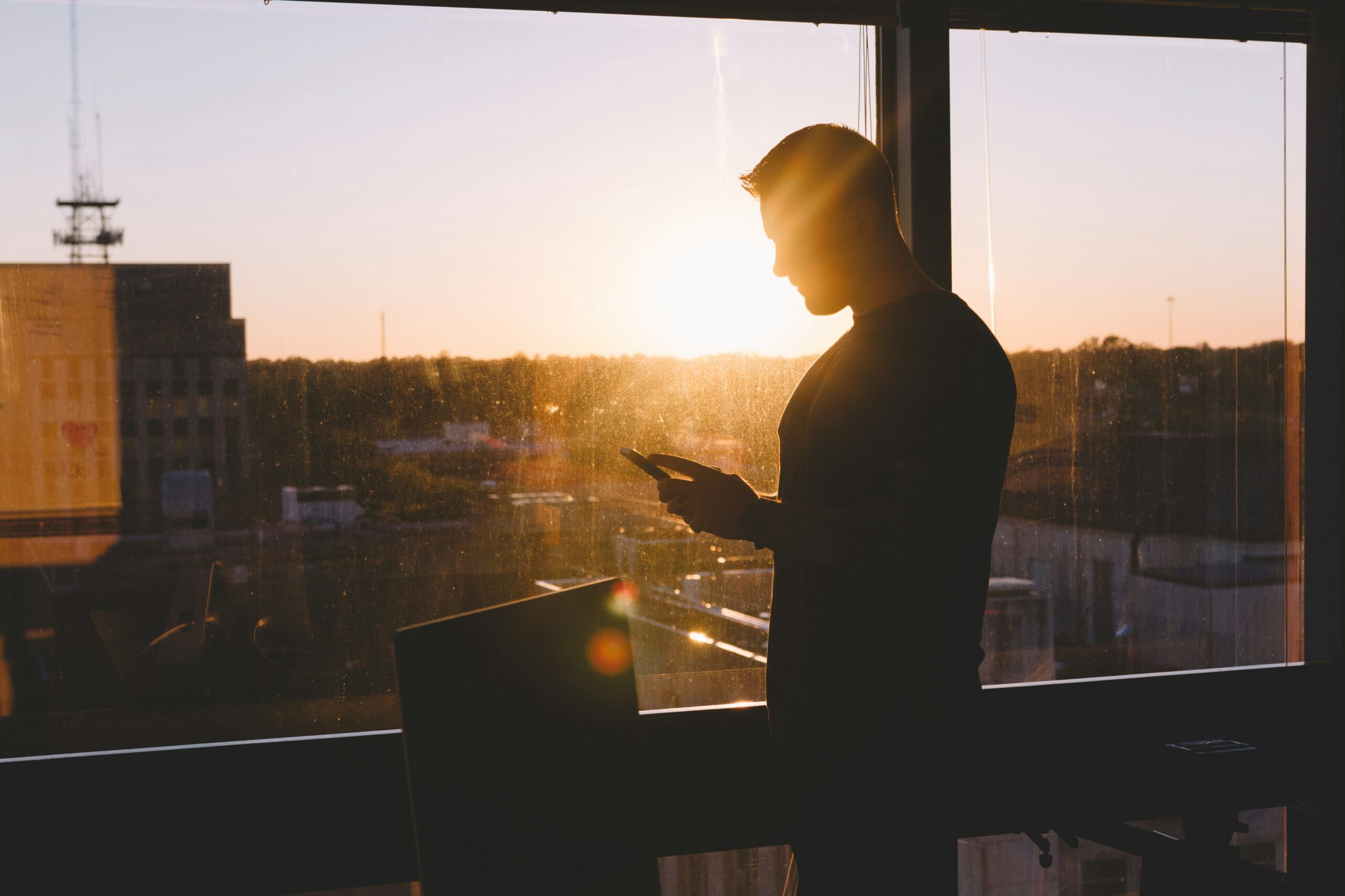 Photo of a silhouette of a man stood in front of office window as sun setting over city.
